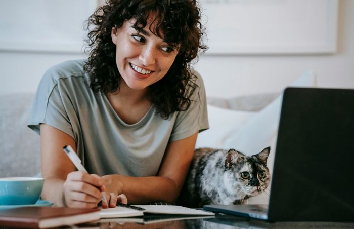 A woman sitting at a table with a laptop and writing in a notebook with a cat next to her preview image